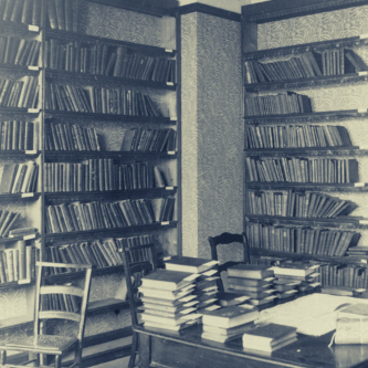 Interior of a library, featuring books on shelves, a desk stacked with books, and two wooden chairs.