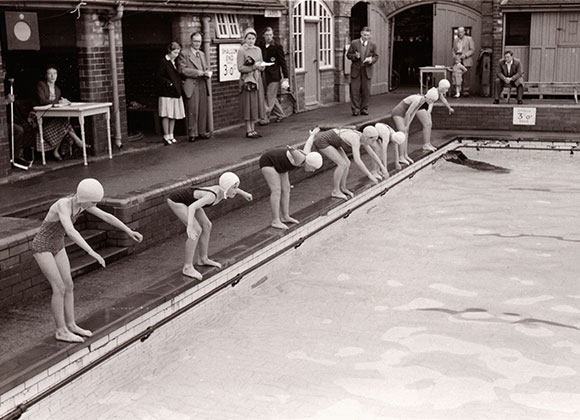 Girls lining up by the edge of a swimming pool in bathing suits and caps ready for a swimming race