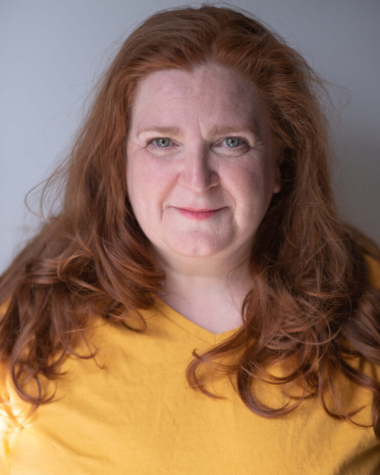 Headshot of woman with long red hair wearing a yellow top in front of a grey background.