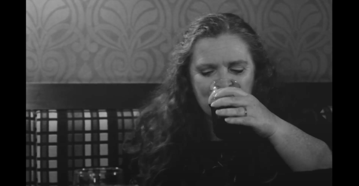 (Black and white) a woman with long hair sat at a pub table drinking a pint.