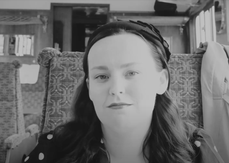 (Black and White) A young woman with long dark hair wearing a polka-dot dress seated at a table on a vintage train.