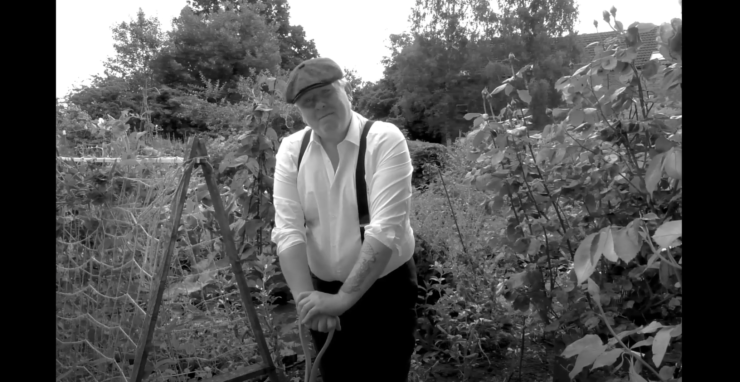 (Black and White) A man wearing a white shirt, braces and a flat cap leans on a space in an allotment.