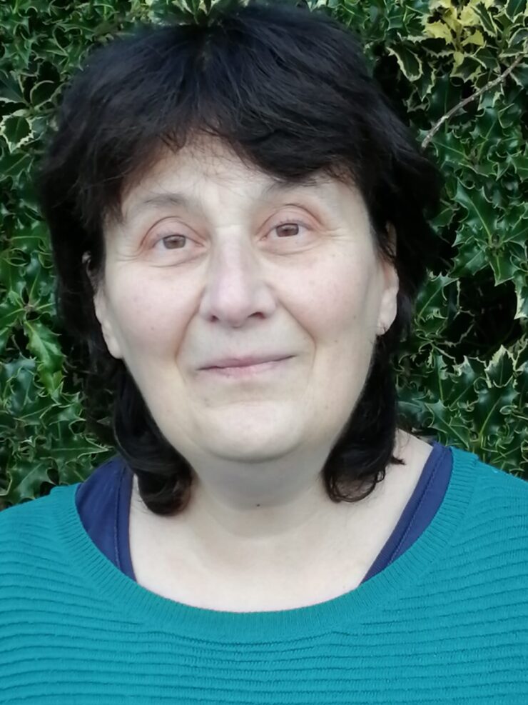 Headshot of woman with shoulder-length black hair pictured outdoors in front of a green hedge.