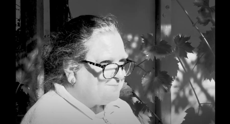 (Black and White) A side-shot close-up view of an older woman with spectacles and her hair tied back. She is sat in a shady corner outdoors with a plant in the background.